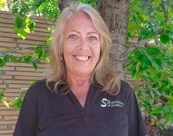 smiling woman with blond long hair in a black shirt in front of a tree