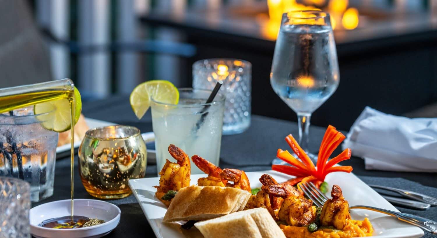 outdoor dining table in front of firepit. Black tablecloth on the table with drinks and white plates with a shrimp dinner