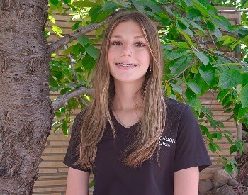 young woman with long brown hair, smiling, in a black shirt in front of a tree