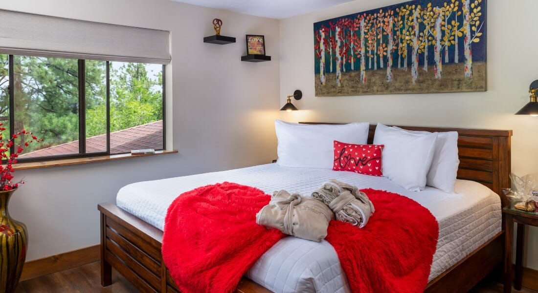 King bed with white linens and red decorative pillow and blanket in front of a contemporary picture of colorful trees and next to an open window in front of trees