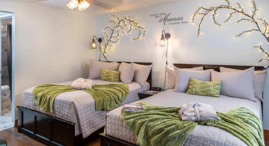 guest room with 2 double beds in gray linens with green decorative pillows and blankets in front of a wall with lighted willow branches