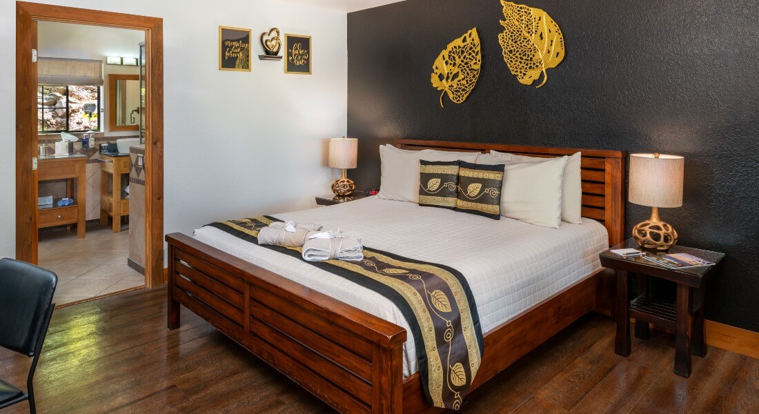 King bed with white linens against a black wall with gold aspen leaves and the door to the bathroom of the room with a window 