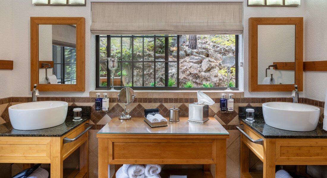 bathroom with countertop with amenities and 2 side vessel sinks with mirrors above them in front of a large window overlooking forested area