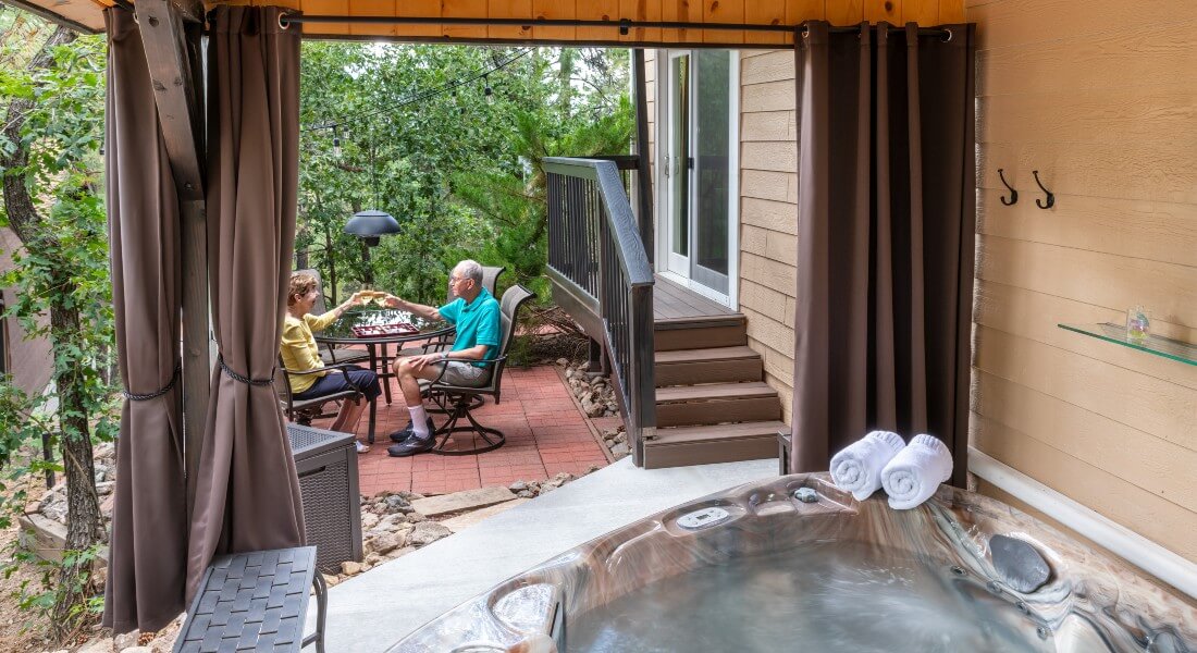 a couple seen in the distance toasting glasses at a glass outdoor table, from the inside of an outdoor hot tub under a gazebo